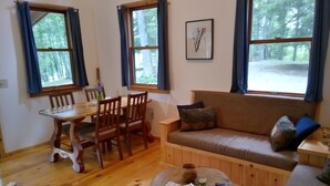 A cozy room with a wooden dining table, chairs, a sofa, and a small coffee table. There are large windows with curtains, a framed picture on the wall, and a view of trees outside. -  Secluded ridge 
 top wilderness retreat with fast wifi and all the comforts  (Wytheville)