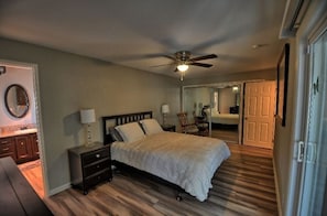 Une chambre à coucher comprenant un lit, des tables de chevet, un ventilateur de plafond, un miroir et une porte donnant sur une autre pièce.