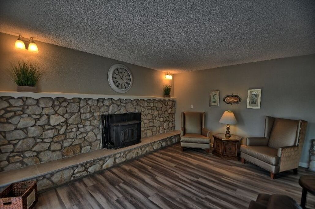 A living room with a stone fireplace, two armchairs, a side table with a lamp, and wall-mounted lights.