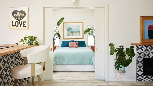 Een moderne slaapkamer met een bed, een bureau en een stoel. Er hangt een schilderij aan de muur en bij het bed staat een bloempot.