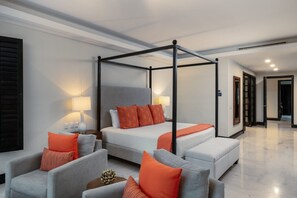 Ein modernes Hotelzimmer mit einem Himmelbett, einer Couch und einem Sessel. Auf dem Nachttisch steht eine Lampe und eine Tür führt in einen weiteren Raum.