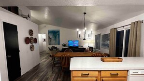 A modern kitchen with wooden cabinets, a dining area with a large table and chairs, and a TV in the background. - Newly Remodeled 3 Bed 2 Bath home, minutes from everything (Pendleton)