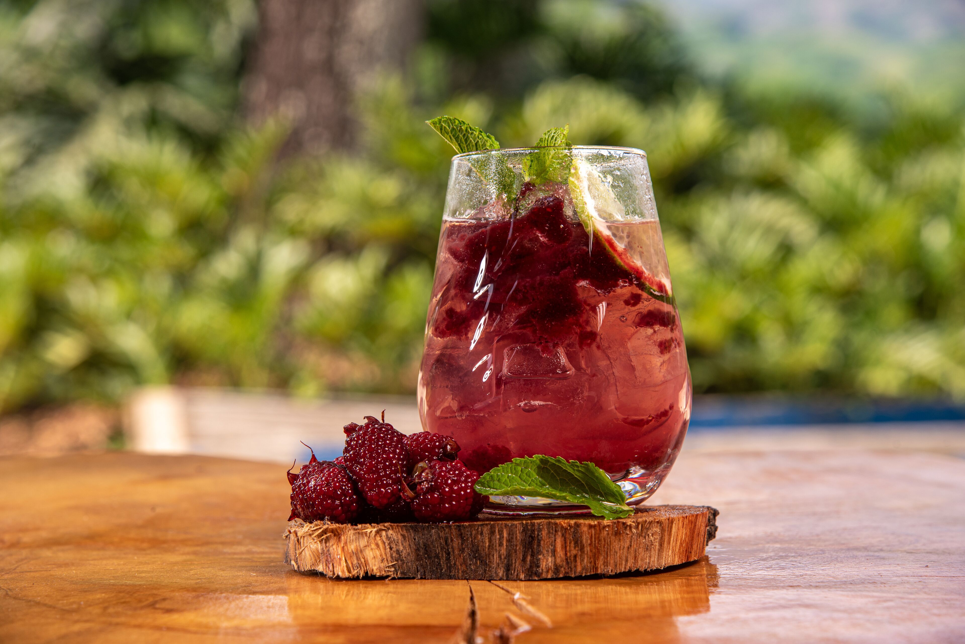 Un vaso de bebida roja con hielo, adornado con hojas de menta, sobre una superficie de madera con frambuesas a un lado.