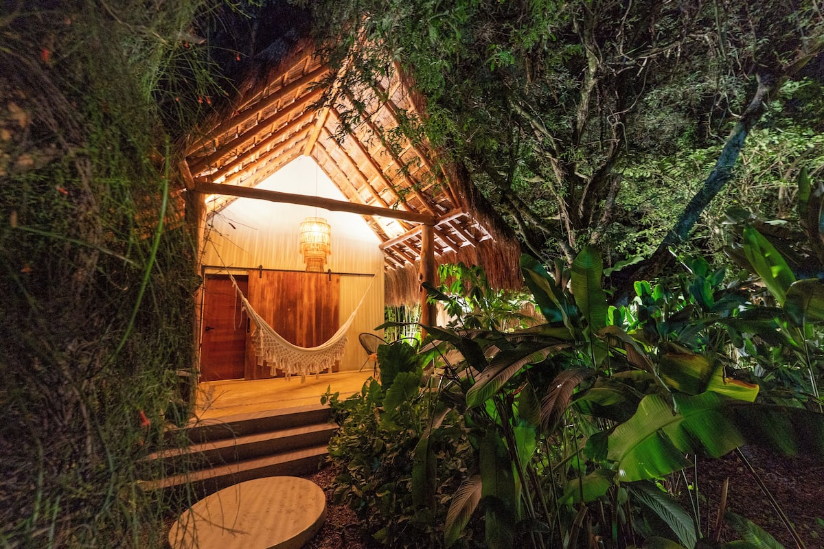 A cozy, thatched-roof structure surrounded by lush greenery, with a hammock and a wooden door leading inside.