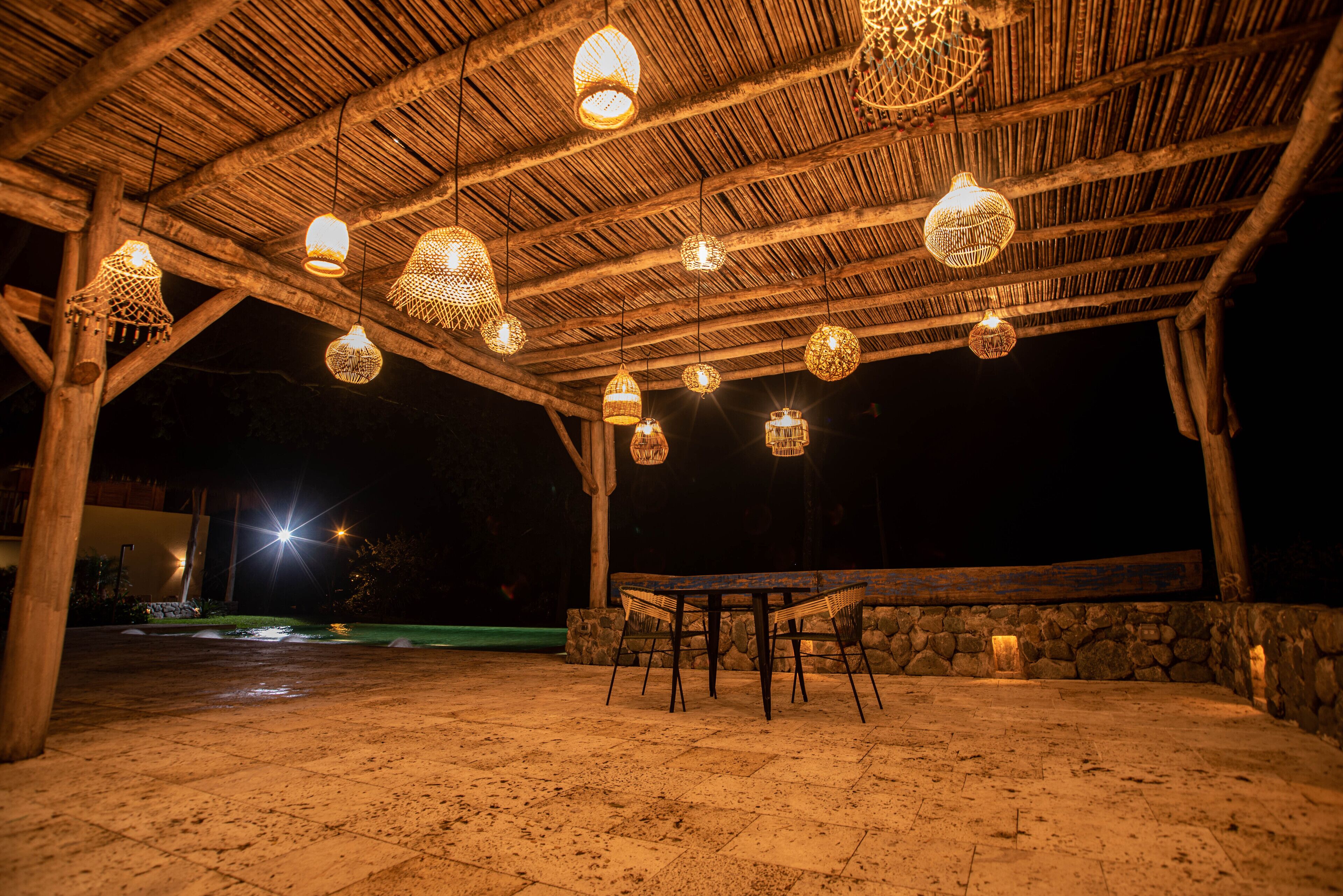 An outdoor covered patio with a rustic wooden ceiling, hanging woven light fixtures, a stone wall, and a table with chairs.