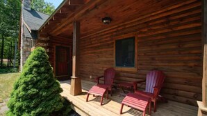 En timmerstuga med en veranda, två Adirondack-stolar och ett litet vintergrönt träd.