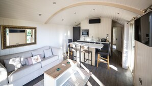 A modern living room with a sofa, coffee table, and a large mirror. A kitchen area with a bar and seating is visible in the background. - Coltsfoot Country Retreat (Knebworth)