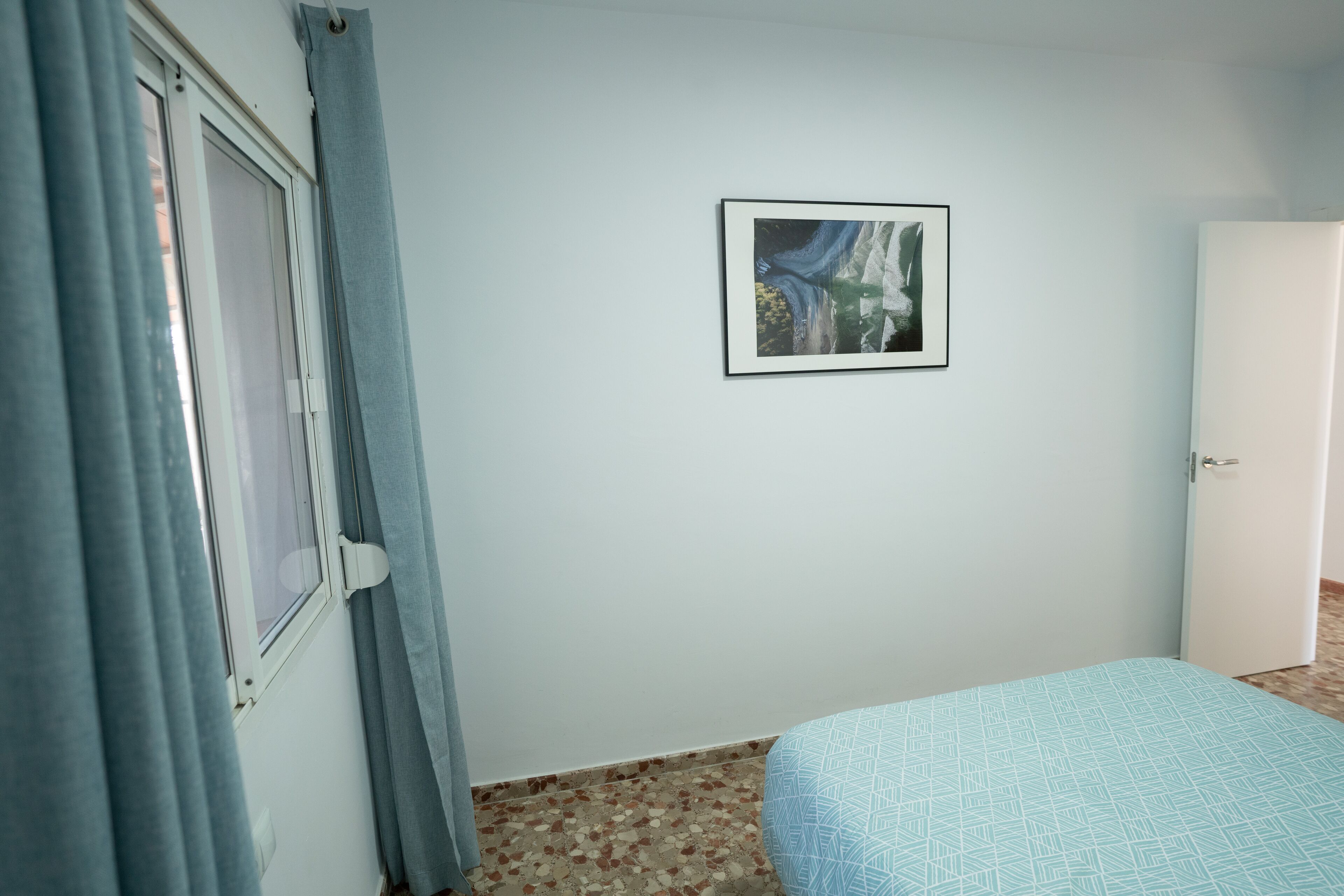 Una habitación con cama, puerta, ventana con cortinas y un cuadro en la pared.