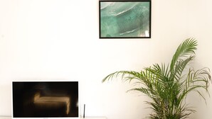 A television on a white stand with a remote control, a framed picture of a wave, and a potted plant. - .................... (AN)