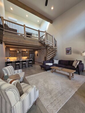Een ruime woonkamer met een hoge plafond, een houten trap, een keuken en een zithoek met een bank en fauteuils.