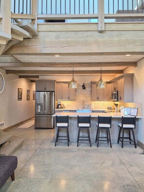 A modern kitchen with a central island, bar stools, stainless steel appliances, pendant lighting, and a view of the outdoors.