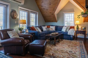 Ein gemütliches Wohnzimmer mit blauem Sofa, Ledersessel und Hocker, Couchtisch aus Holz und einem gemusterten Teppich.