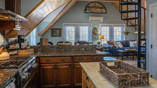 A kitchen with a stone countertop, wooden cabinets, and a gas stove. There is a dining area with a table and chairs, and a loft bedroom with a wooden staircase.