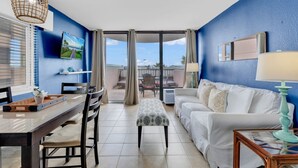 A room with a dining table, chairs, a white sofa, a small table with a lamp, and a large window with curtains. - Palms Resort Getaway (Myrtle Beach)