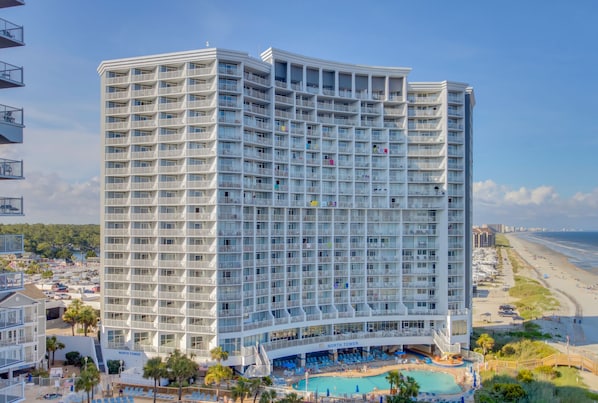 A multi-story hotel with balconies, a pool area, and a view of the beach.