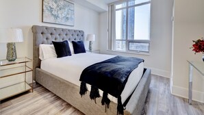 Une chambre moderne avec une tête de lit capitonnée, une grande fenêtre donnant sur la ville, une table d’appoint en verre et une lampe de chevet.