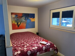 A bedroom with a bed, a window with blinds, a painting on the wall, and a view of the outside.