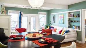 Un coin repas avec une table ronde, des chaises et un lustre. On aperçoit un canapé blanc et une bibliothèque à l’arrière-plan.