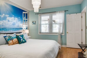 Une chambre avec un grand lit, une fenêtre ornée de rideaux légers, une porte blanche et une fresque murale représentant une scène de plage.