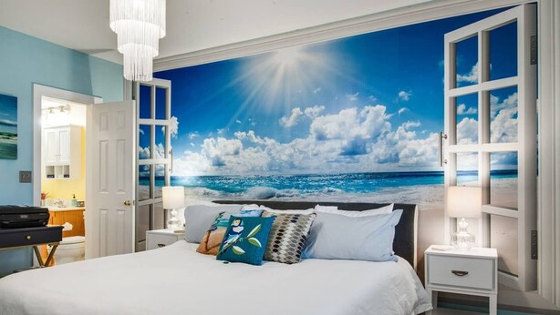 Une chambre avec un grand lit, une table de chevet, une lampe et une vue sur l’océan par une fenêtre ouverte.