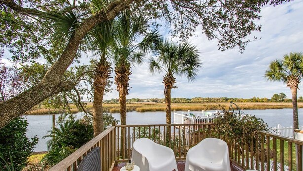 A deck with white chairs and a red cushion, overlooking a lake with palm trees and a white fence. - Waterfront Fishing Dock FirePit HotTub Deck GrillB (Hanahan)
