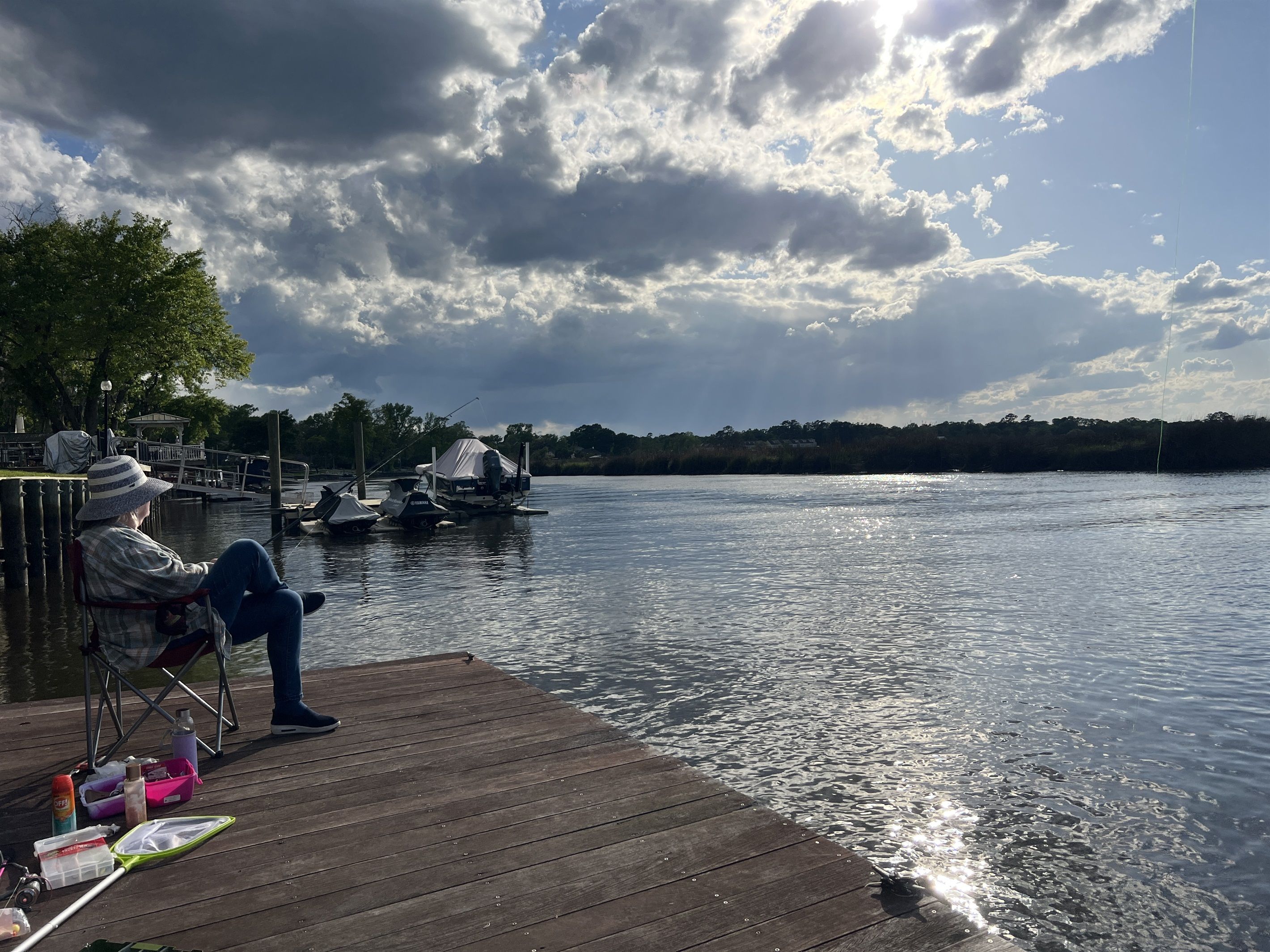 A wooden dock with a person seated, a fishing rod, and boats on the water.