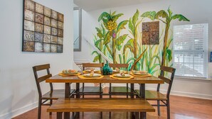 A dining area with a wooden table set for four, surrounded by chairs. A large mural of tropical plants is on the wall behind the table. - Waterfront Fishing Dock FirePit HotTub Deck GrillB (Hanahan)