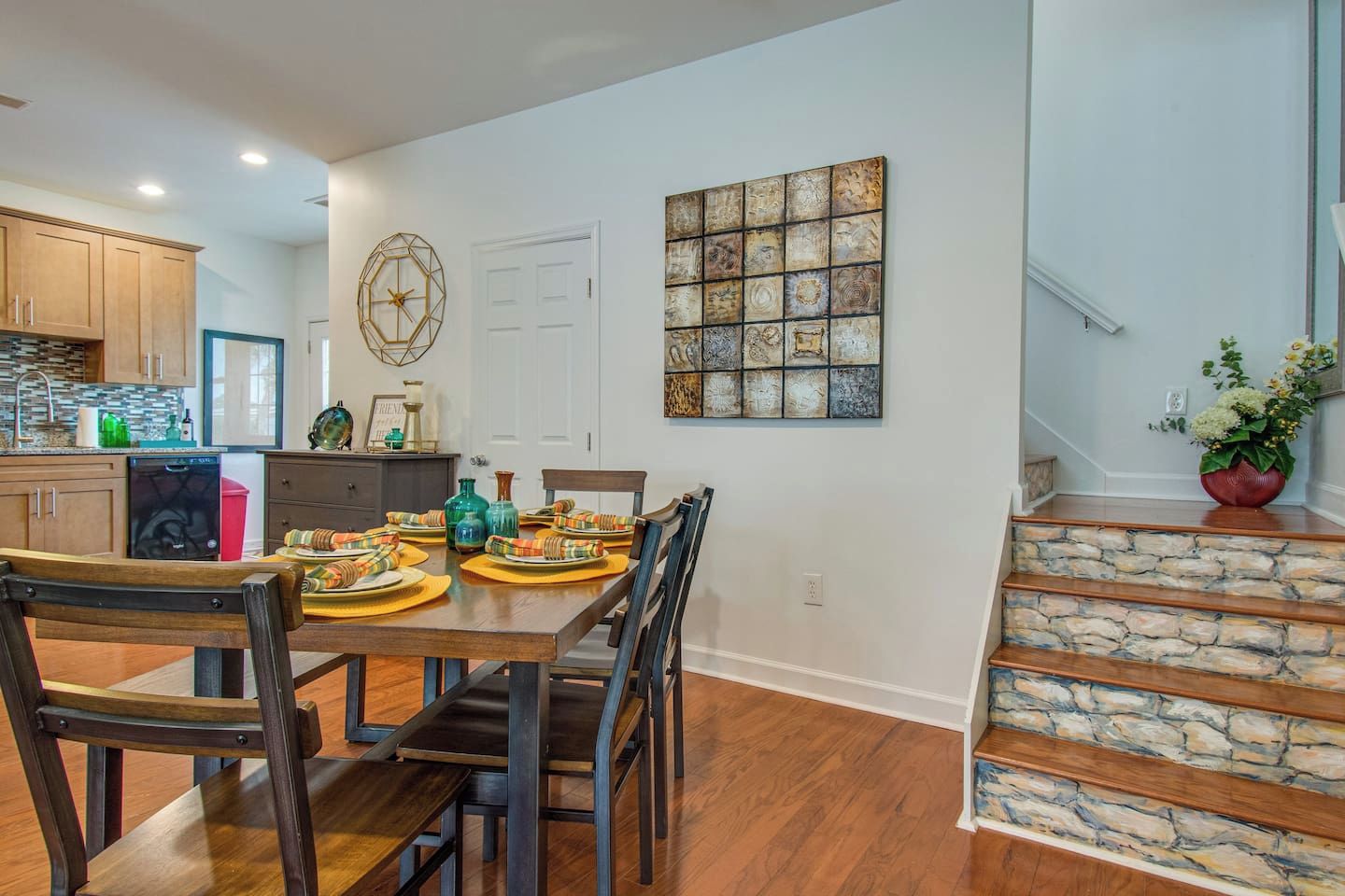 A dining area with a wooden table set for four, surrounded by chairs. A kitchen is visible in the background with wooden cabinets and a dishwasher.