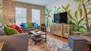 A living room with a stone wall, a flat-screen TV, a sofa with colorful cushions, a coffee table, and a patterned rug. - Waterfront Fishing Dock FirePit HotTub Deck GrillB (Hanahan)