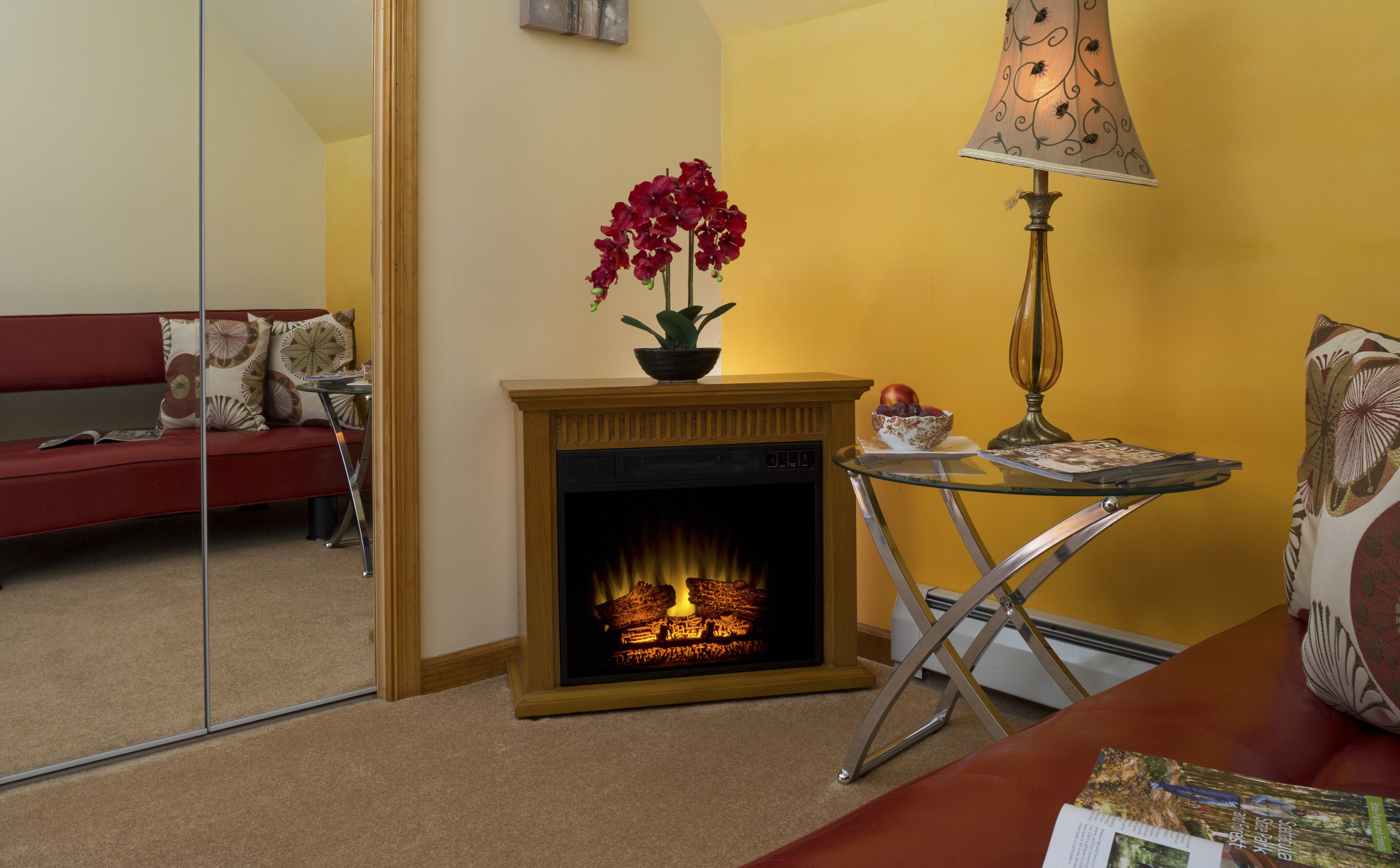A cozy living room with a fireplace, a red sofa, a small table with a lamp, and a mirror reflecting the room.