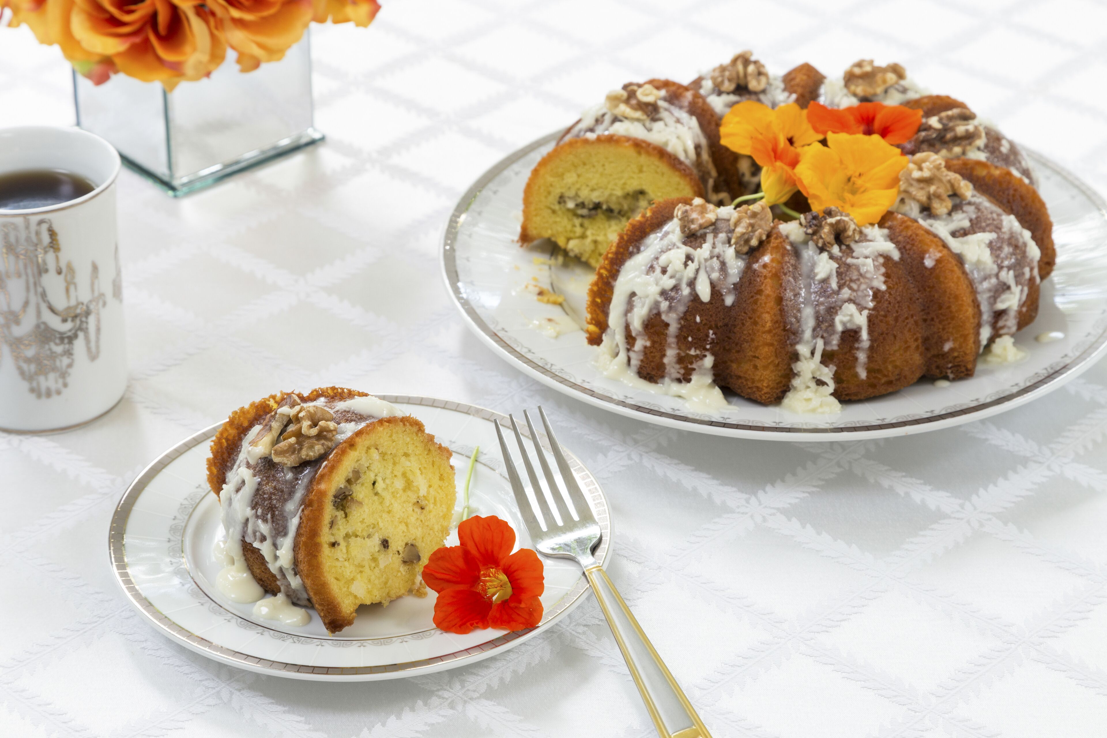 Un pastel Bundt con crema y nueces sobre un plato, una porción más pequeña en un plato más pequeño, y un tenedor con una flor.