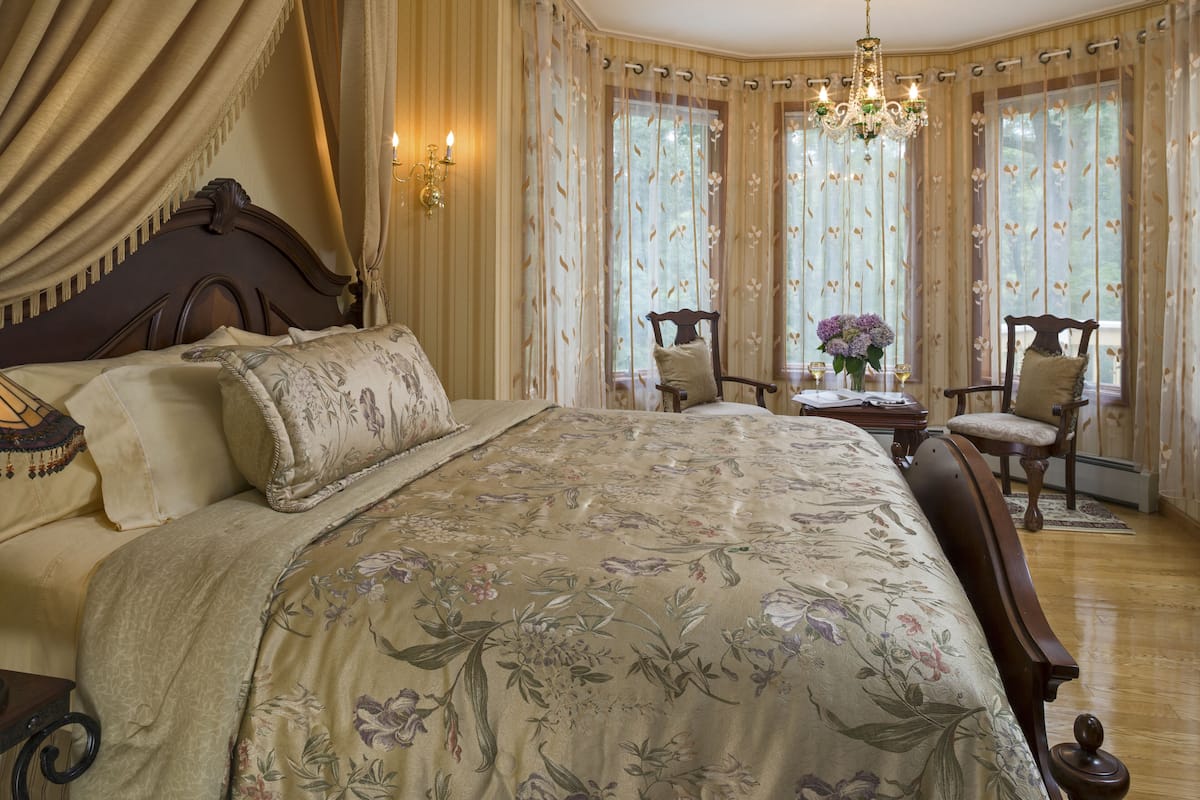 A bedroom with a large bed, floral bedspread, two chairs, a table with a vase, and a chandelier.