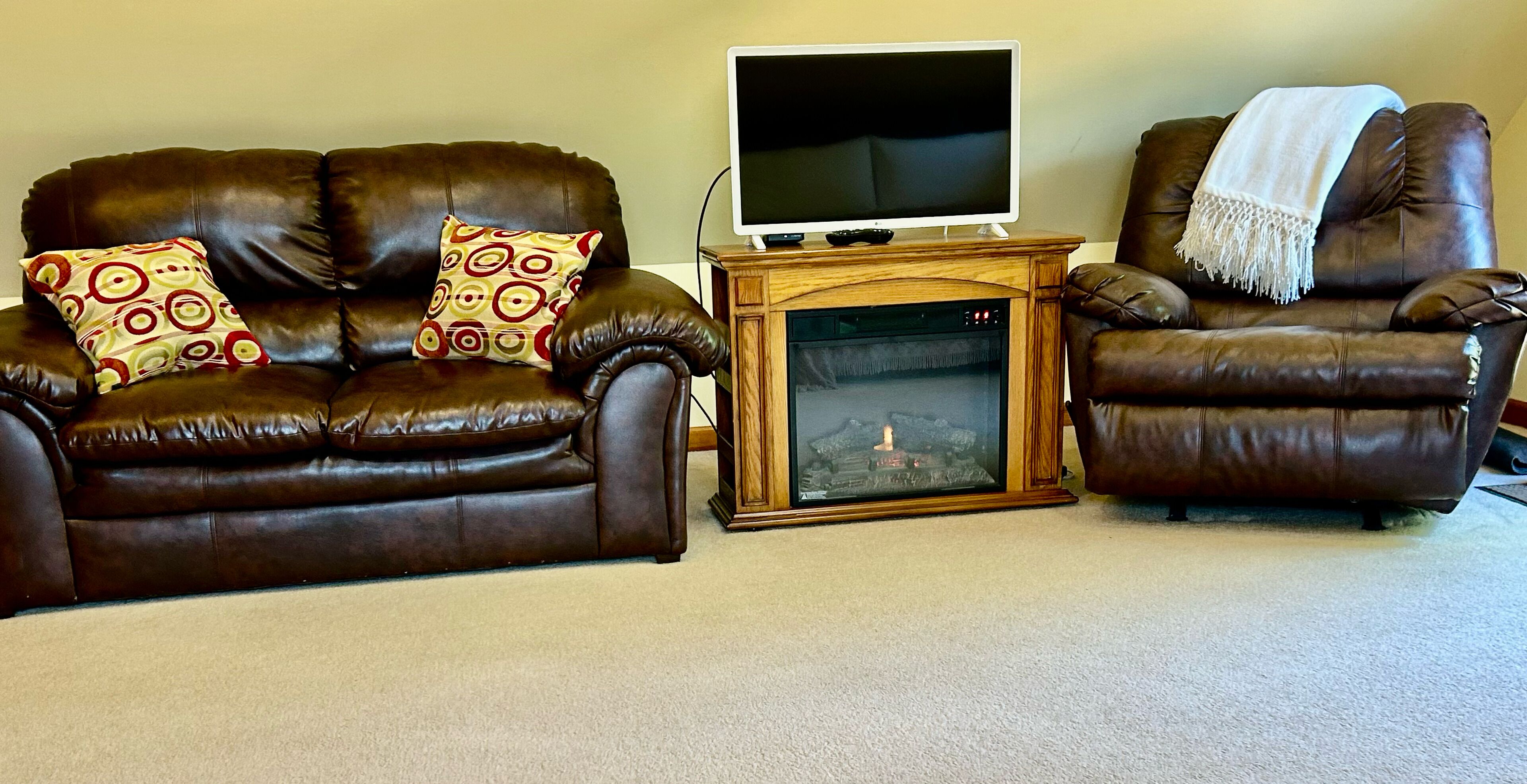 A living room with a brown leather sofa, two patterned pillows, a wooden entertainment unit with a TV, and a fireplace.