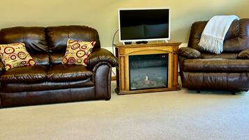 A living room with a brown leather sofa, two patterned pillows, a wooden entertainment unit with a TV, and a fireplace.