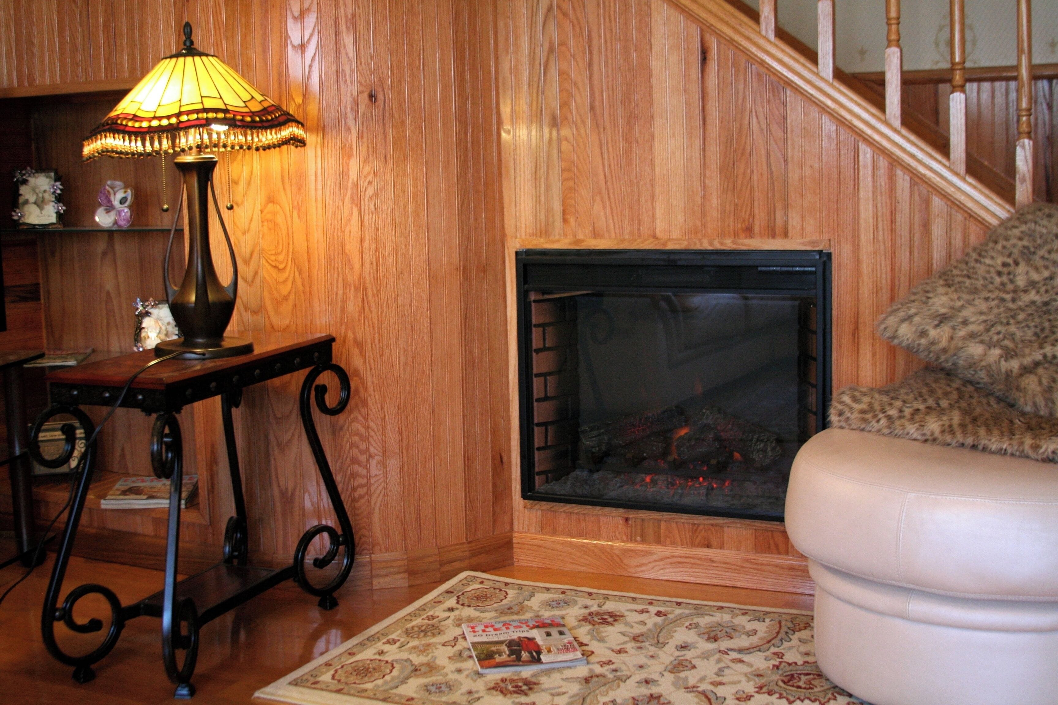 A wooden interior with a fireplace, a table with a lamp, and a patterned rug.