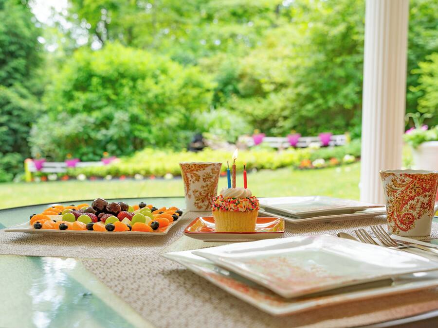 Une table dressée avec un plateau de fruits, un cupcake décoré de bougies et une tasse ornée de motifs floraux, avec une vue sur un jardin en arrière-plan.