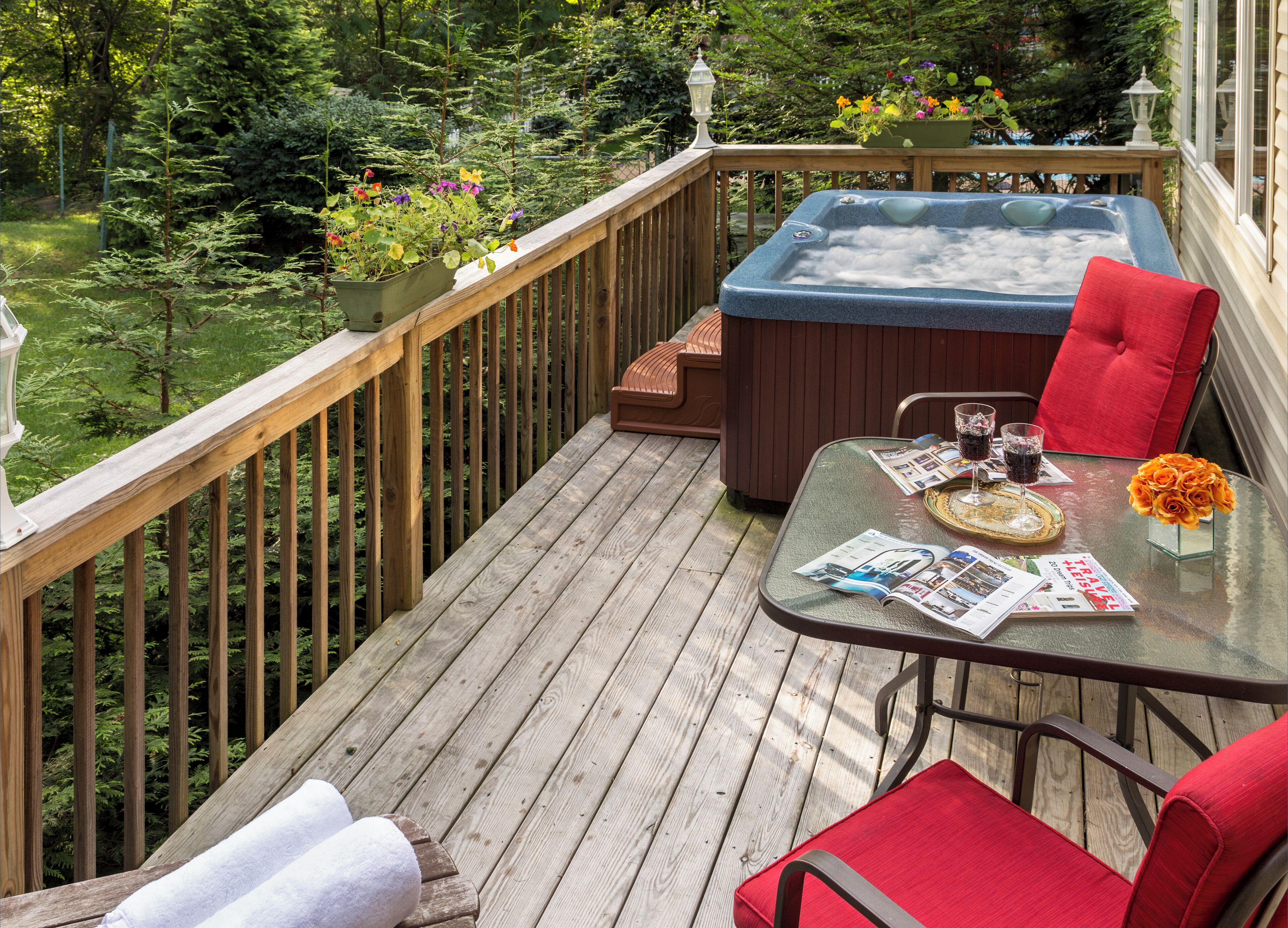 A wooden deck with a hot tub, red chairs, and a glass table with magazines and drinks.