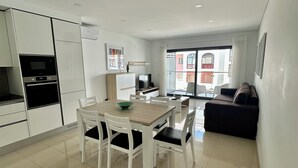 A modern kitchen with white cabinets, a dining table with chairs, a black sofa, and a large window with a view of buildings. - Apartment with 2 bedrooms 100 meters from the beach / New (Quarteira)