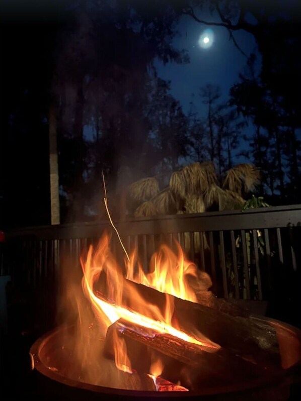Ein Lagerfeuer mit Flammen, umgeben von einer Holzterrasse und Bäumen, unter einem Nachthimmel mit Mond.