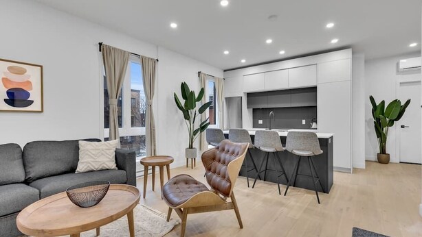 A modern living room with a grey sofa, a wooden coffee table, and a leather armchair. There is a dining area with a grey table and chairs, and a kitchen with white cabinets and a stainless steel sink. - Sunny, bright & natural apt, centrally located (Montréal)