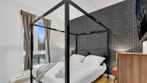 A modern bedroom with a four-poster bed, a wooden dresser, and a cityscape view through the window. - Sunny, bright & natural apt, centrally located (Montréal)
