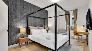 A modern bedroom with a four-poster bed, a geometric-patterned wall, a small wooden side table, a desk with a chair, and a large window with curtains. - Spacious & peaceful 2 bedrooms, sleep 6 (Montréal)