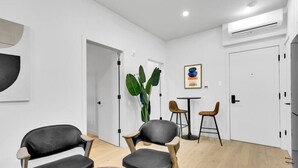 A modern living room with two black leather chairs, a wooden coffee table, and a small round table with two stools. There is a large green plant in the corner and a framed artwork on the wall.