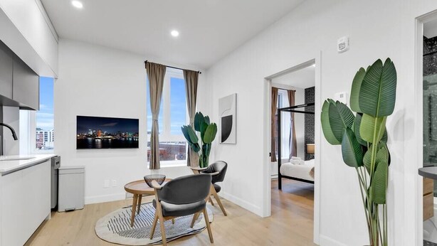 A modern apartment with a kitchen, dining area, and living space. It features a large window with a city view, a dining table with chairs, and a potted plant.