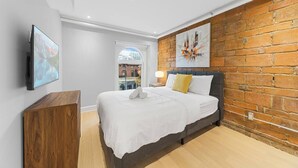A modern hotel room with a large bed, a wooden nightstand, a flat-screen TV, and a brick accent wall. - Stunning 2 beds, 2 baths, AAA location, rooftop (Montréal)