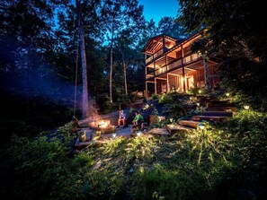 A two-story wooden cabin with a deck, surrounded by forest. There is a fire pit with people sitting around it. - Red Apple Cabin 🍎3 Bedrooms, 3 Baths, Amazing views Mountaintop 15 MIN DOWNTOWN (Cherry Log)