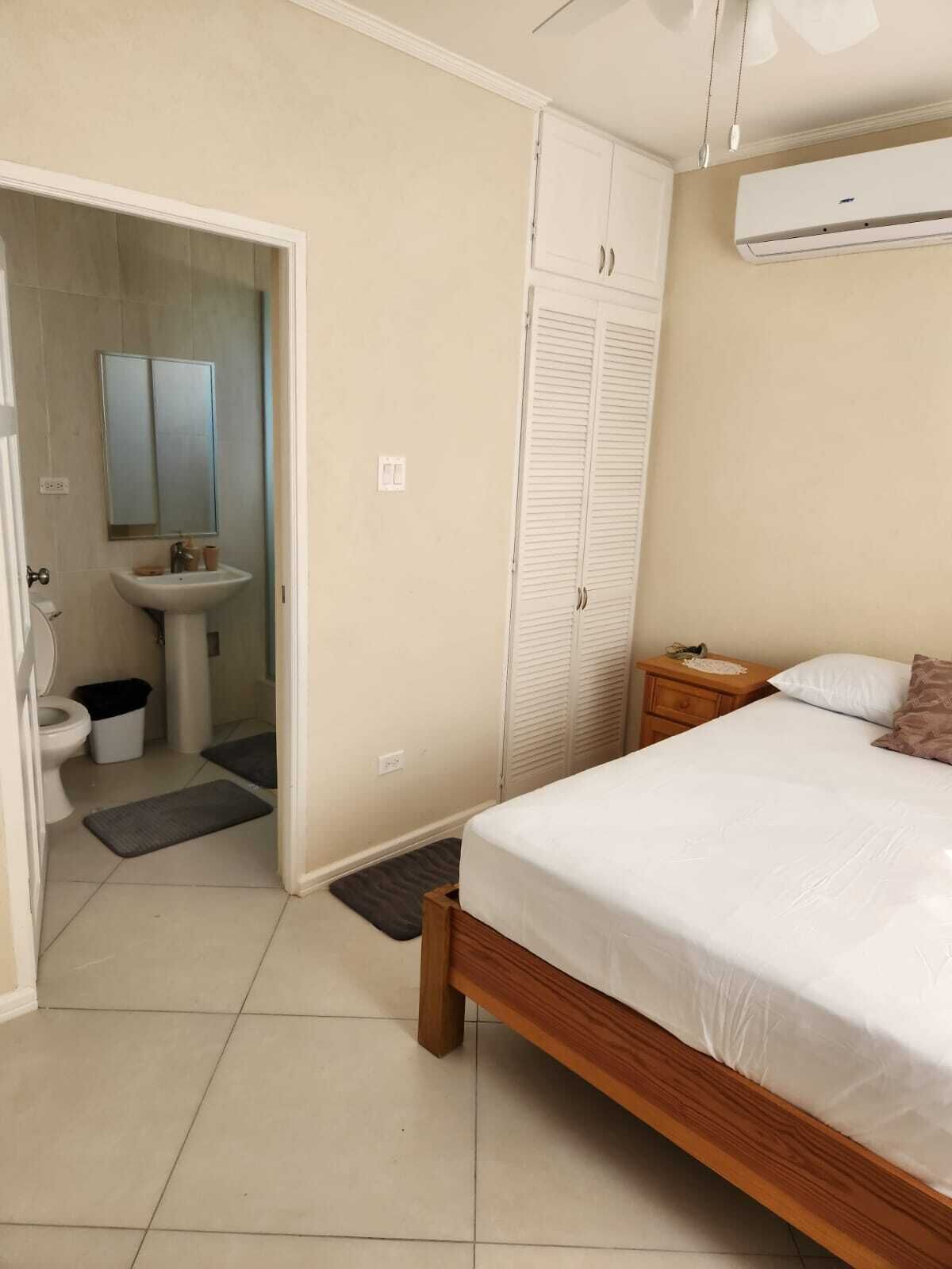 A bedroom with a wooden bed, a bedside table, a nightstand, a white wall-mounted air conditioner, a wardrobe, and a bathroom with a sink and mirror.