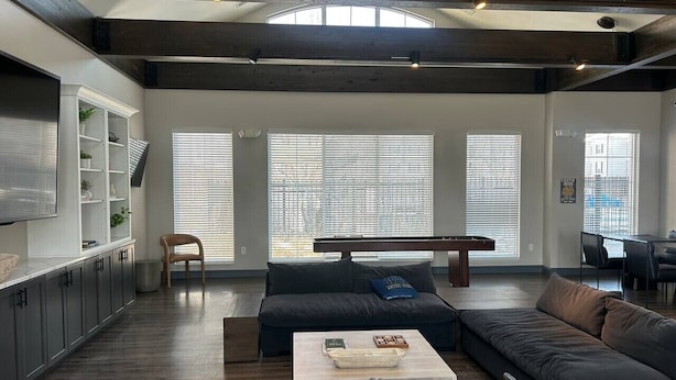 A spacious living area with a dark-colored sofa, a coffee table, and a pool table. There is a dining area with a table and chairs, and a kitchen area with cabinets and a countertop.