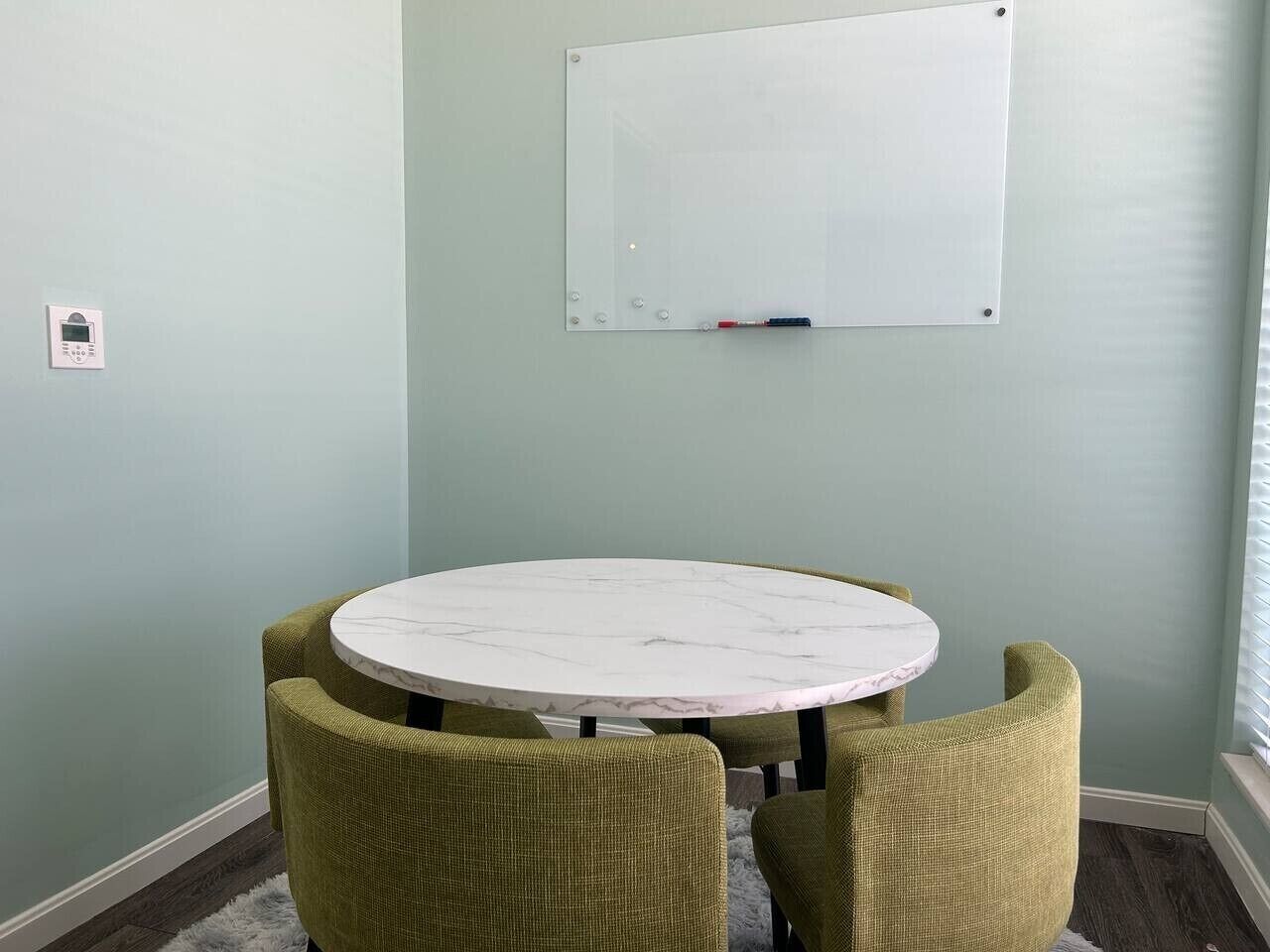 A room with a round table, four chairs, a whiteboard, and a thermostat.