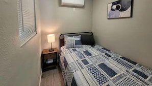 Un dormitorio pequeño y ordenado con una cama individual, una mesita de noche con lámpara, aire acondicionado de pared y un cuadro enmarcado.